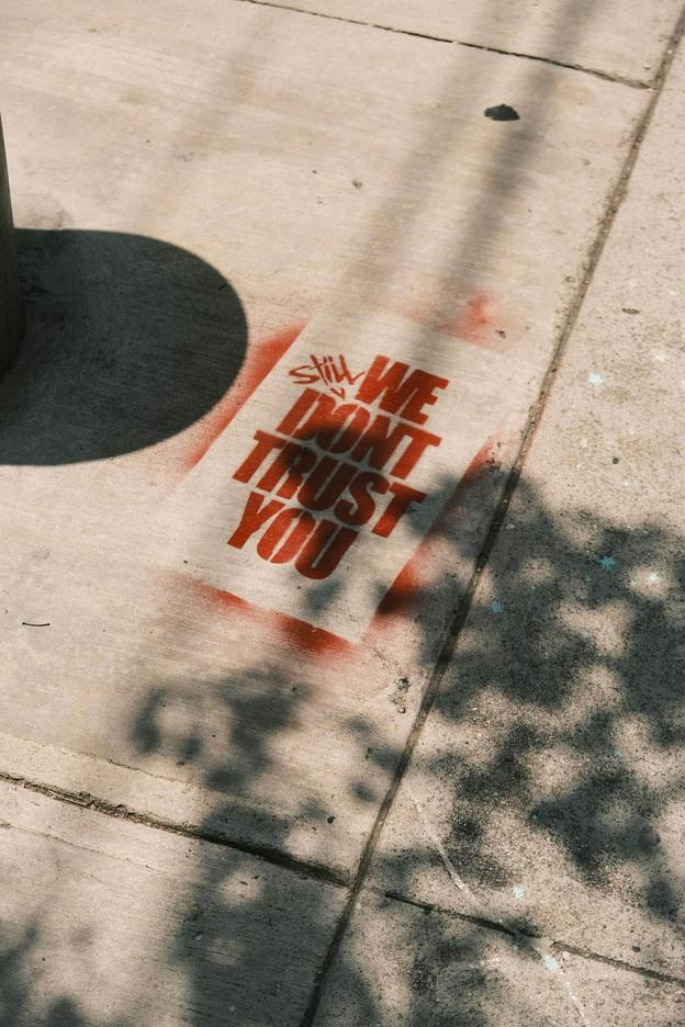 A red spray-painted stencil on a sunlit sidewalk reading "still we don't trust you"
